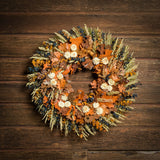 A dried wreath made of preserved spiral eucalyptus in amber and navy blue colors with a mix of sudan grass, preserved oak leaves in orange and cafe colors, and cream strawflower accents hung on a dark wood background.