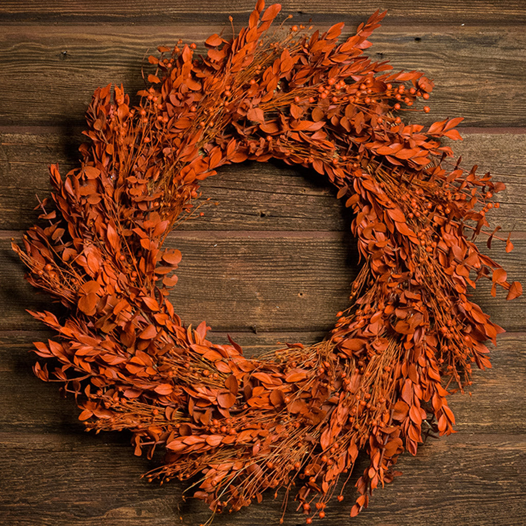 A close-up of a deep orange wreath made of preserved orange myrtle, preserved eucalyptus, and orange flax pods hanging on a wood background. 