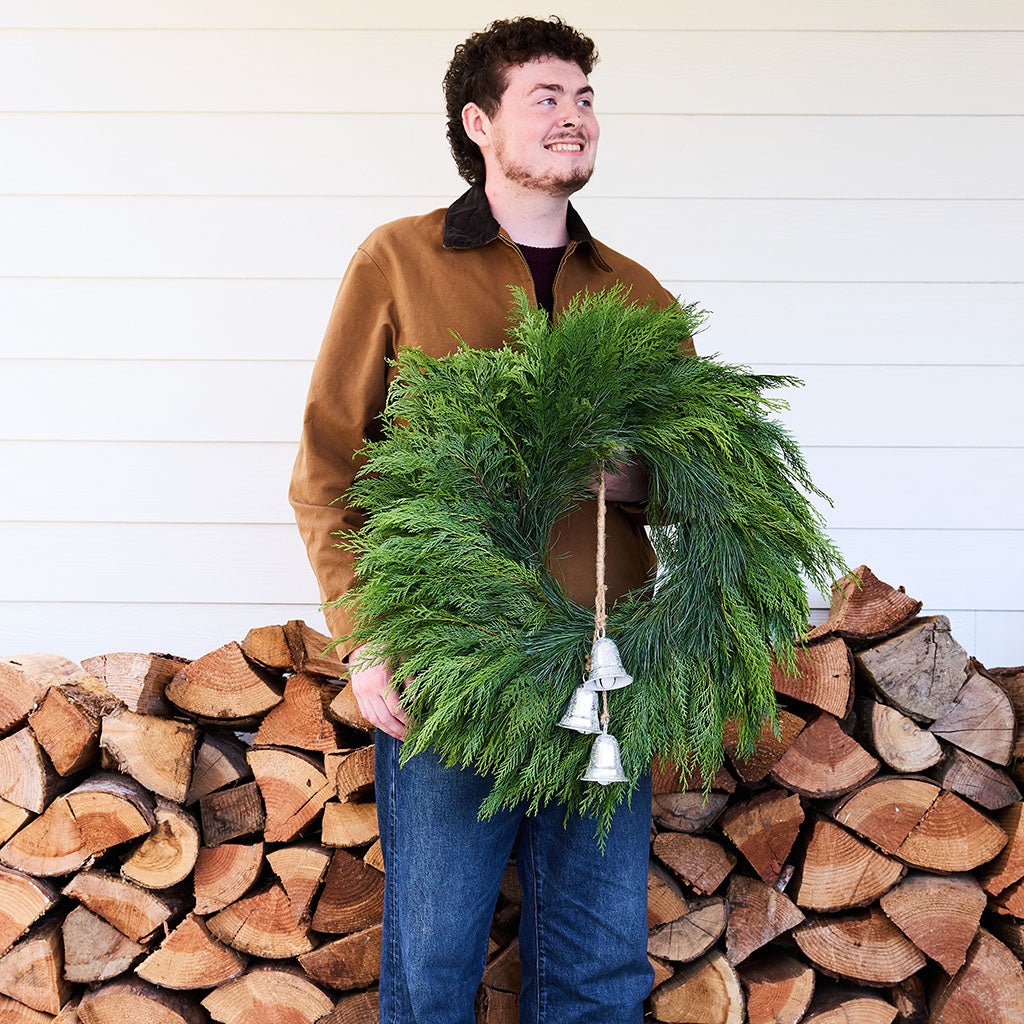 A man wearing a brown jacket and blue jeans stands in front of a white siding wall and a stacked firewood pile, holding a green evergreen wreath with small silver bells.