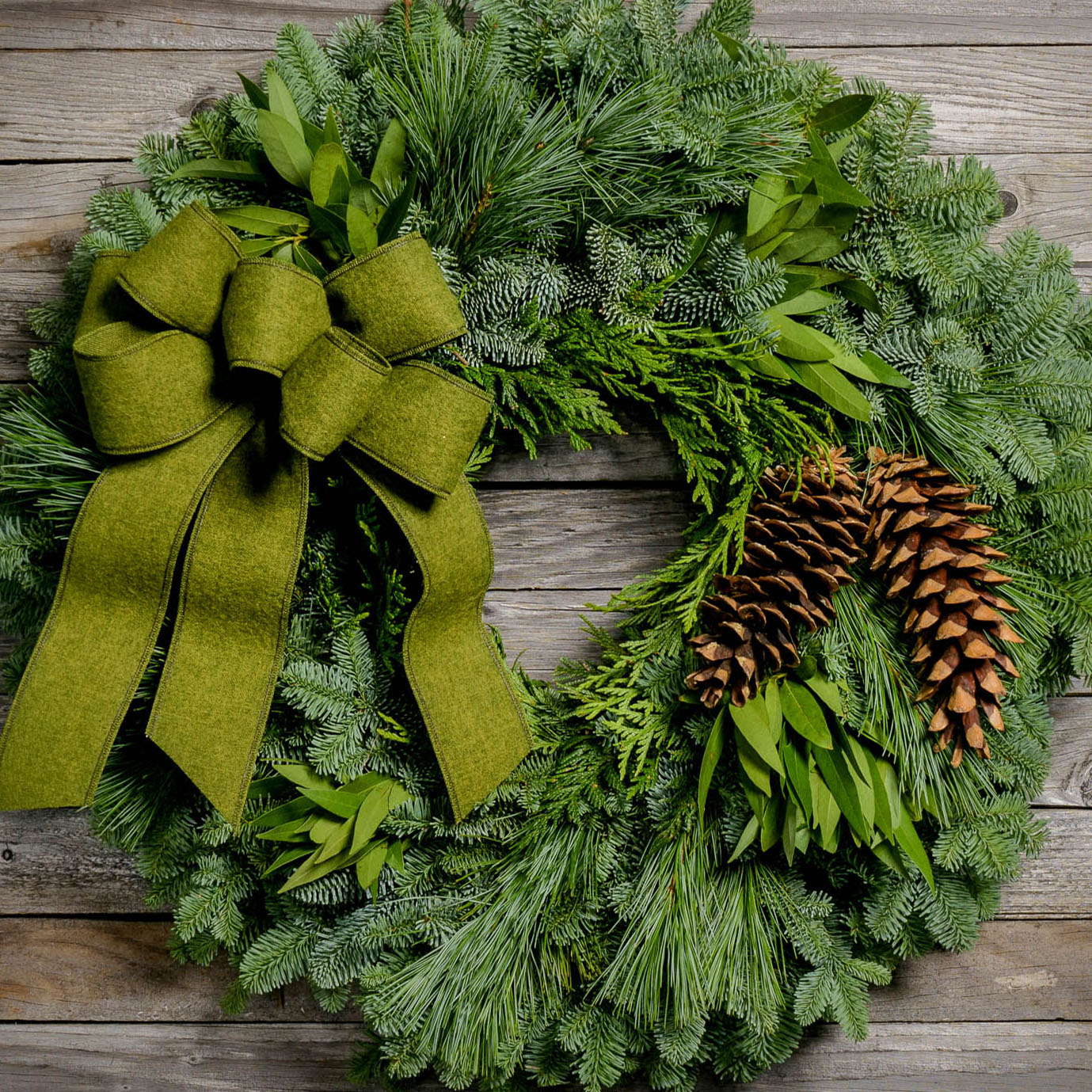 Christmas wreath with bay leaves, pine cones with a moss green brushed linen bow on a light wood background.