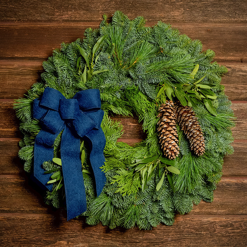 Christmas wreath with bay leaves, pine cones with a blue brushed linen bow on a dark wood background.