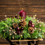 Centerpiece made of  verdant fresh noble fir, white pine, and incense cedar decorated with glittery beads and shiny burgundy balls, natural pinecones and a deep burgundy pillar candle on a small wood table.