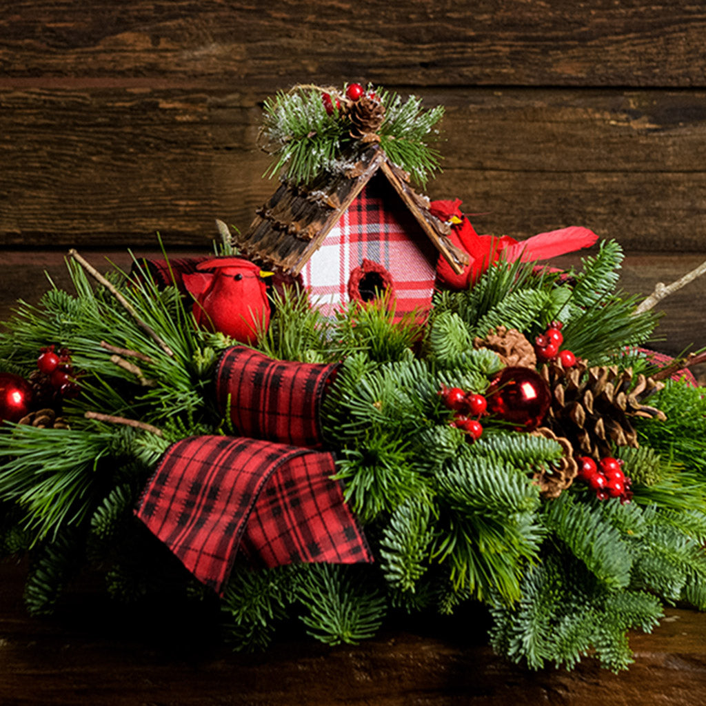 A holiday centerpiece of noble fir, white pine, incense cedar, natural pinecone, red berry & red ball clusters, 2 cardinal birds, natural stick clusters, a plaid birdhouse, and 2 tartan bows with a wood background. 