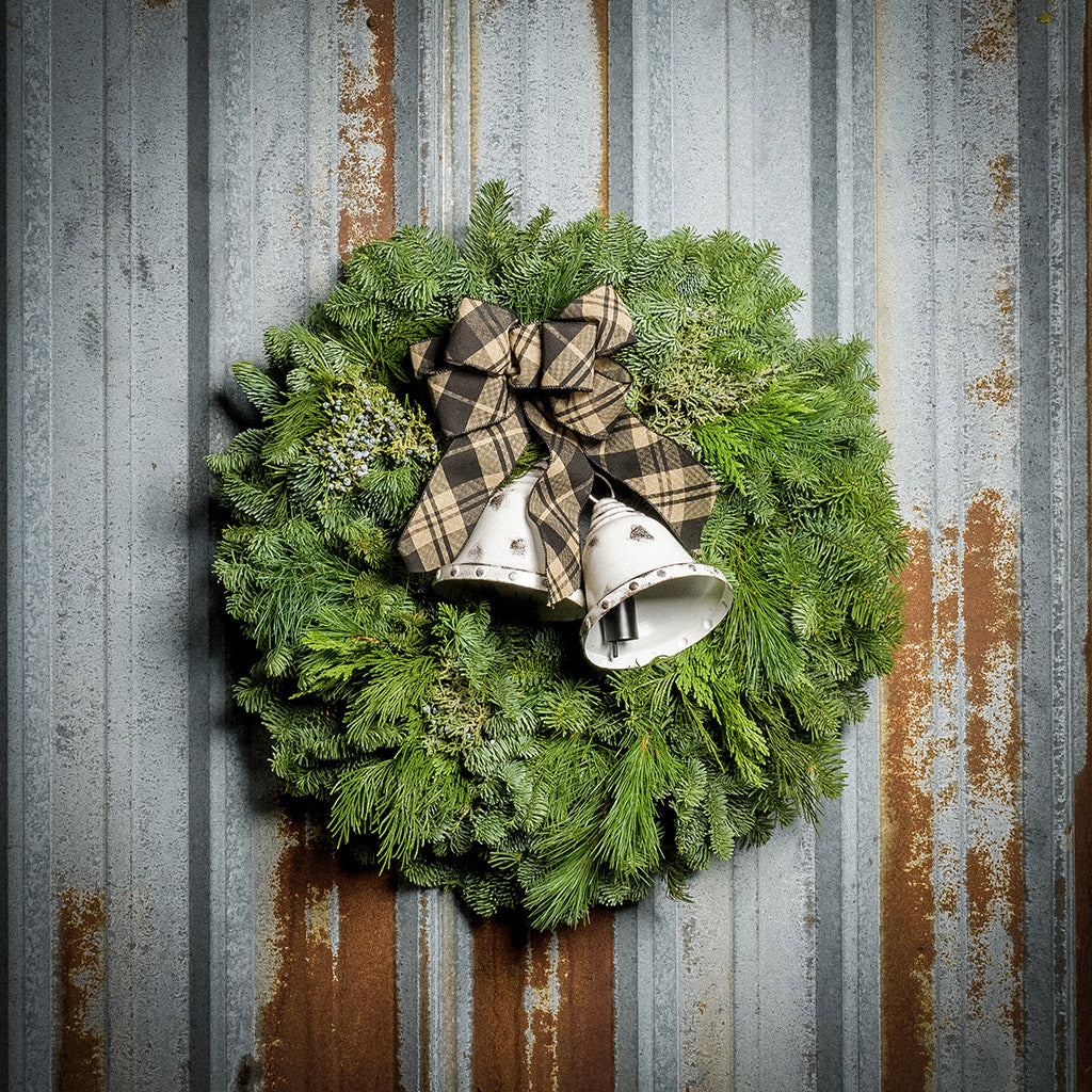 A wreath made of noble fir, incense cedar, juniper, and white pine with white metal bells, and a black and tan plaid linen bow on a rustic metal background.
