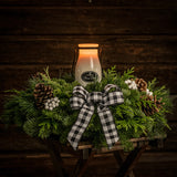 An arrangement made of noble fir, incense cedar, and white pine with Austrian pinecones, white berry clusters, a black and white gingham linen bow, and a milk bottle candle with an evergreen fir scent with a dark wooden background.
