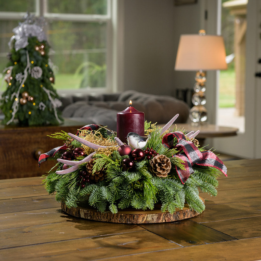 A holiday centerpiece of noble fir, white pine, incense cedar, 2 chickadee birds, 2 red and black plaid bows, 4 burgundy ball clusters, 6 Australian pinecones, 6 burgundy berry clusters, moss, 2 faux antlers, and a burgundy pillar candle sitting on a wood round on a counter. 