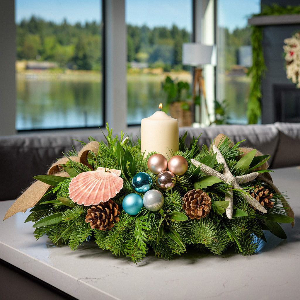 An arrangement made of noble fir, white pine, incense cedar, and bay leaves, with a seashell, seastar, aqua ball clusters, champagne ball clusters, Austrian pinecones, an ivory pillar candle, and tan linen ribbons sitting on a counter.
