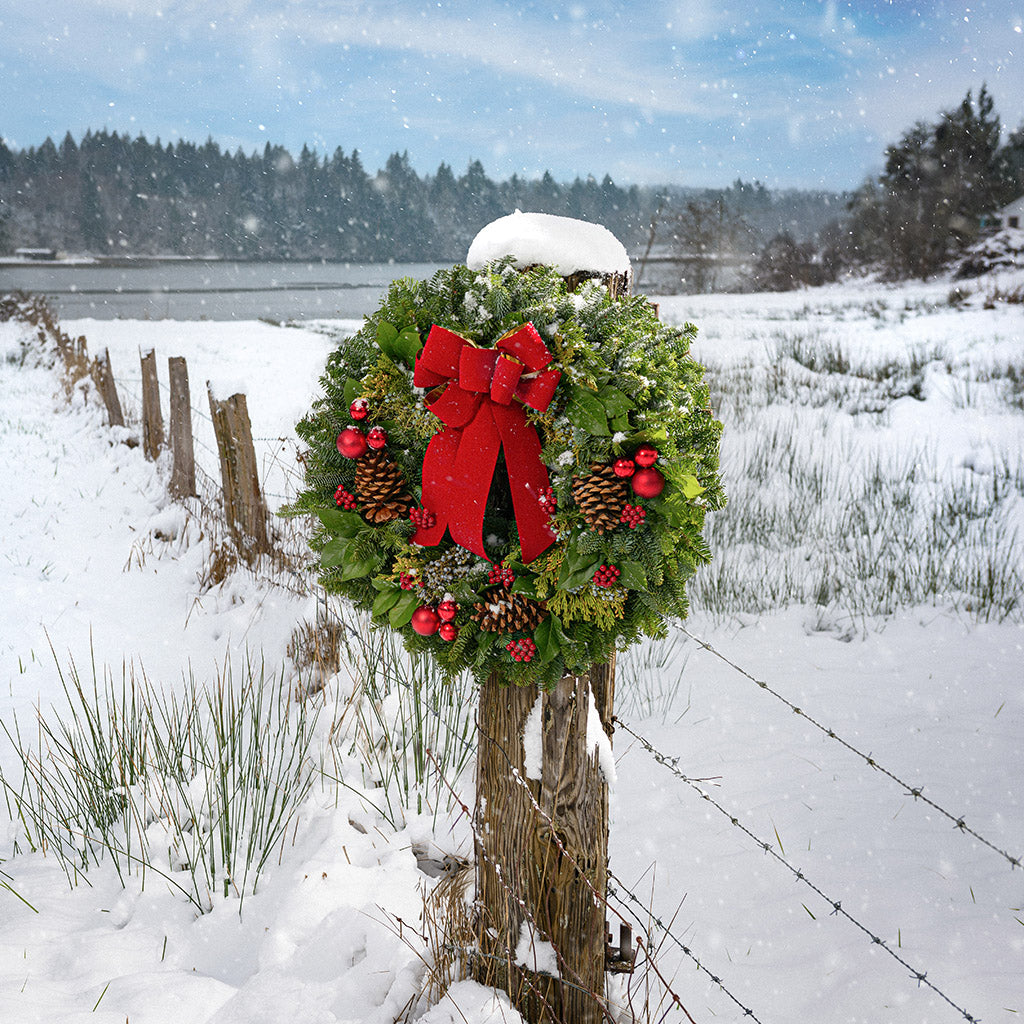 Holiday wreath made of noble fir, incense cedar, juniper, and salal leaves with ponderosa pine cones, faux red berry clusters, ball clusters, and a red bow hanging on a post in the snow