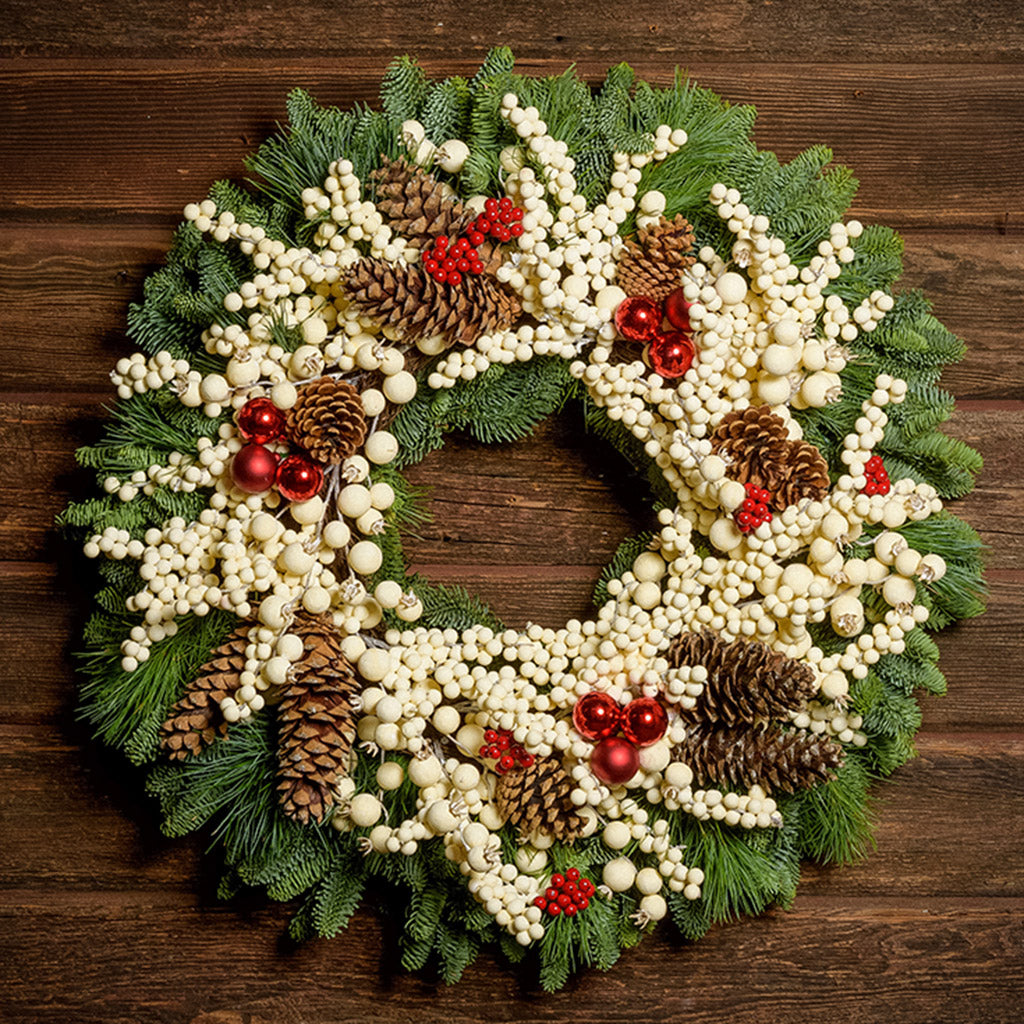 A wreath made of noble fir and pine with cream-colored berries, Australian pinecones, red ball ornaments, and red berry clusters on a dark wooden background.