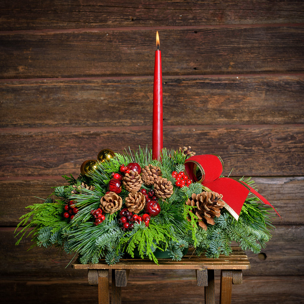 Christmas centerpiece with pine cones, gold balls and red berries with red velveteen bows and a red taper candle with a dark wood background.