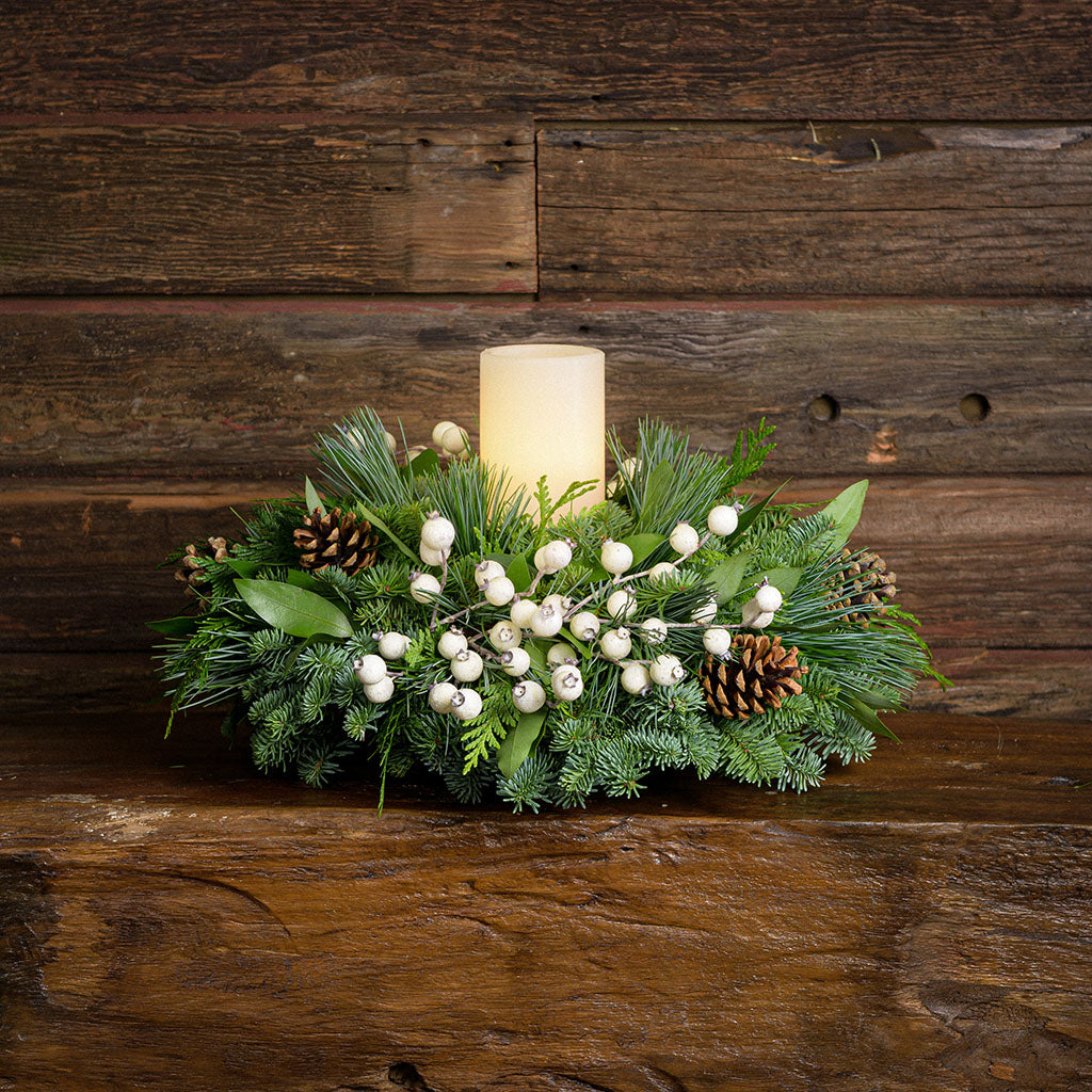 Centerpiece made of noble fir, pine, cedar and bay leaf with white berry clusters, Australian pine cones and 1 white LED pillar candle on a wooden table against a wood wall
