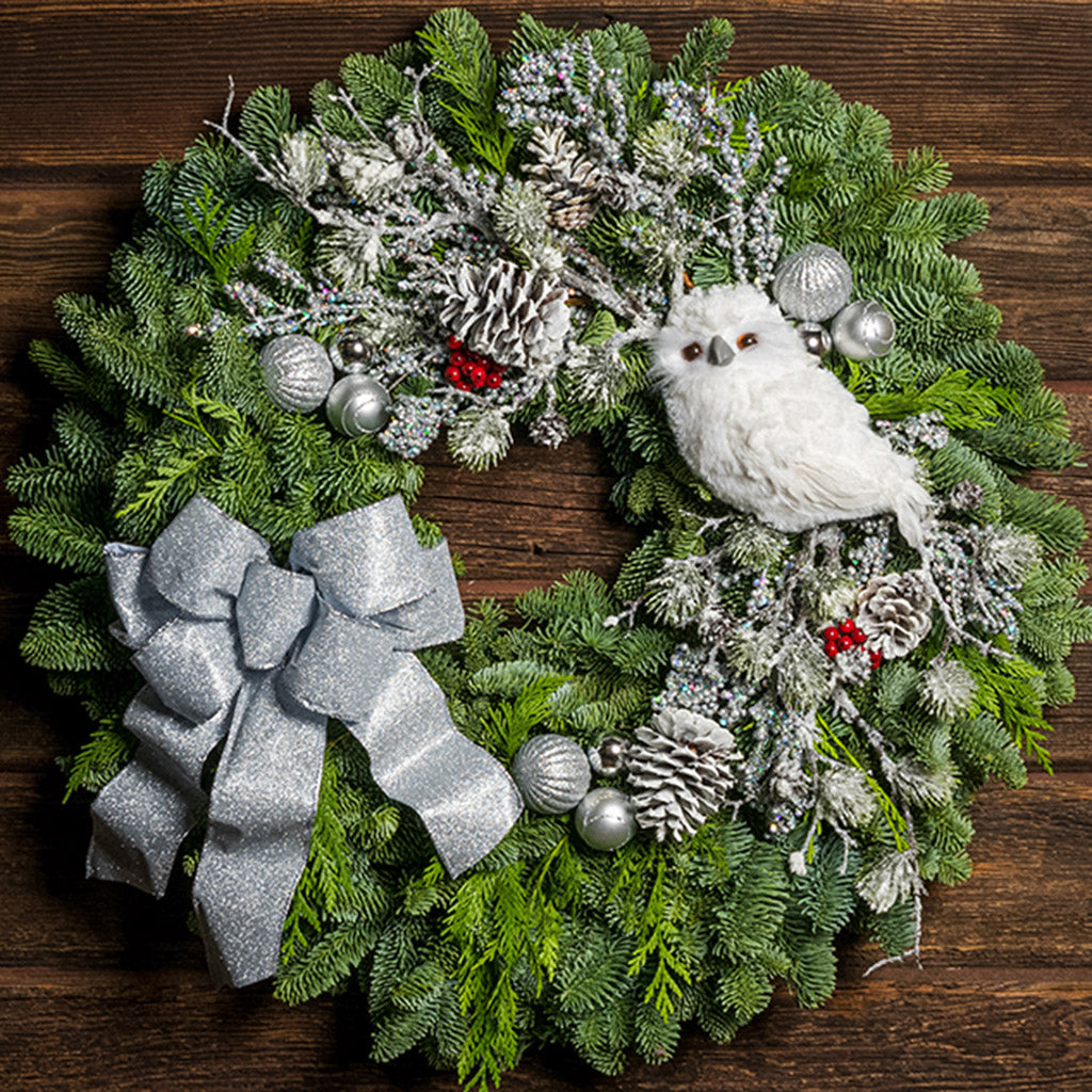 Holiday wreath of noble fir and western red cedar with a snowy white owl, faux flocked branch, small glitter fans, frosted branches, red berry clusters, shiny silver ball clusters, pinecones, and a silver glitter bow on a wood background.