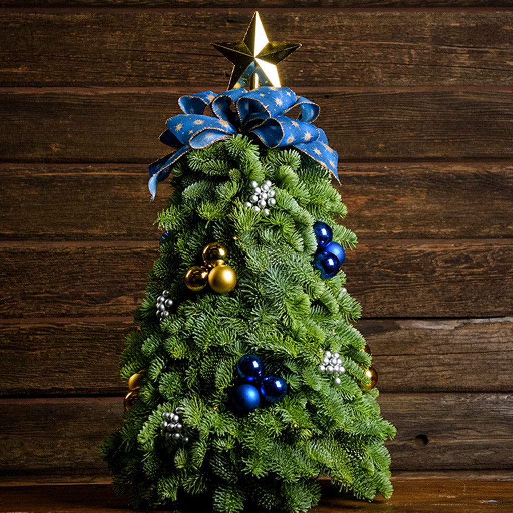 A table top tree made of noble fir with 4 gold ball clusters, 4 blue ball clusters, 8 silver berry clusters, a gold star tree-topper, and a blue starry bow with a wood background. 