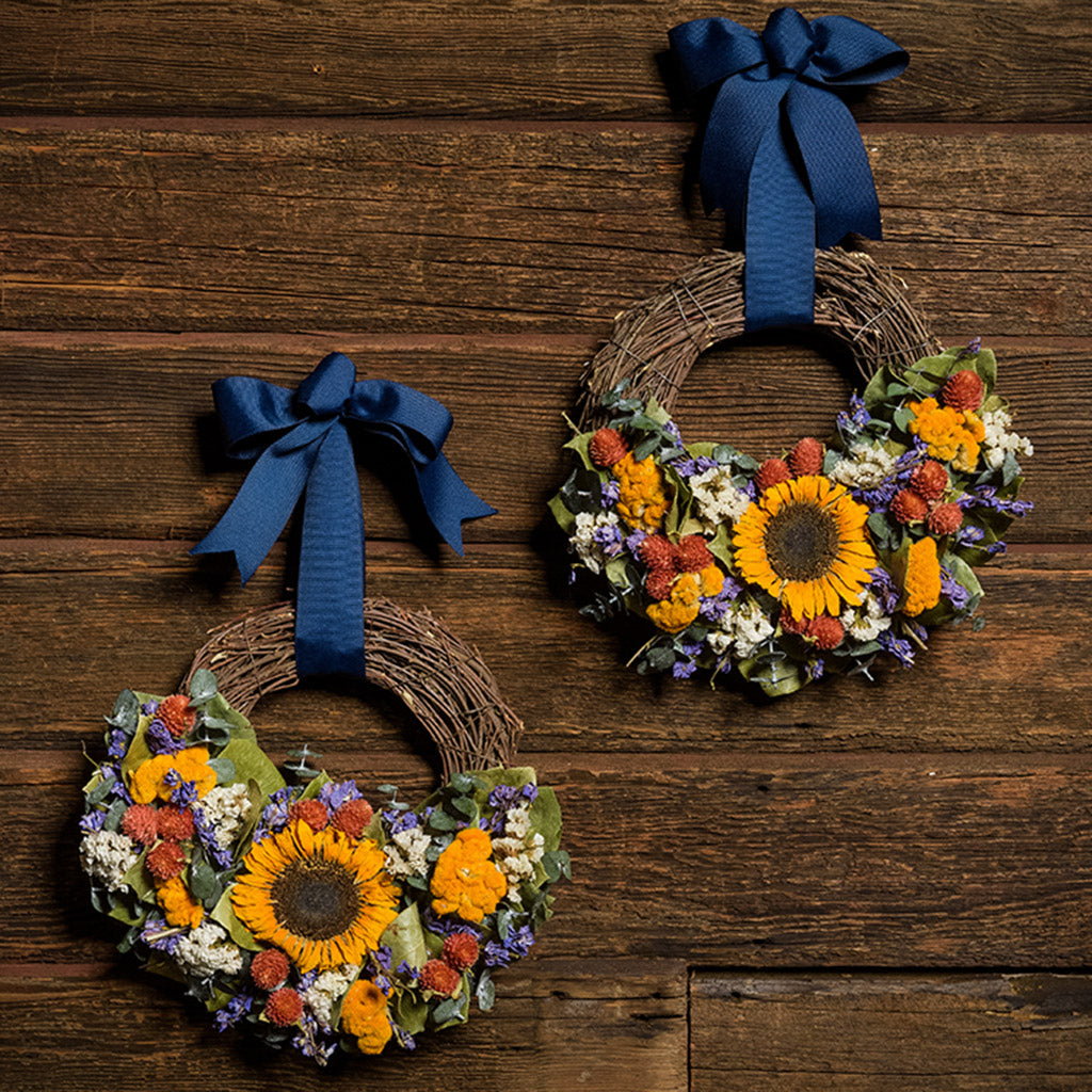 Two 10" wreaths made of eucalyptus, yellow coxcomb, white statice, red globe amaranthus, natural salal leaves, and real sunflowers hung on a dark wood background with grosgrain navy-blue bows.