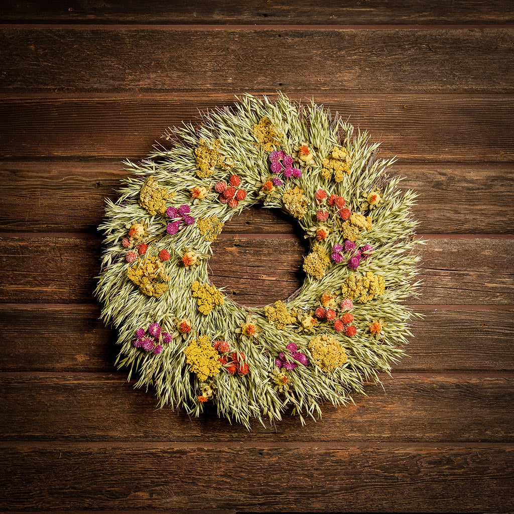 A wreath made of All-natural dried yarrow, natural oats, red and purple globe amaranthus, and safflowers on a dark wood background.