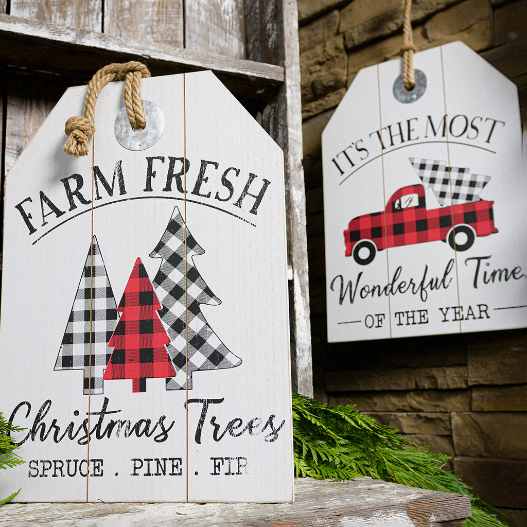 Sturdy wood signs hanging from a thick rustic jute rope. Both have beautiful rustic white-washed coloring. One sign says "Farm Fresh Christmas Trees - Spruce, Pine, Fir" with charming buffalo-plaid trees. The other sign says "It's the Most Wonderful Time of the Year" with a buffalo-plaid pickup truck bringing home a Christmas tree.