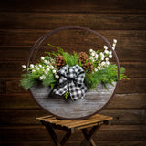An arrangement made of noble fir, Western red cedar, and white pine with ponderosa pinecones, white mini pomegranate branches, and a black and white gingham linen bow in a wood and metal hanging container with a dark wood background.