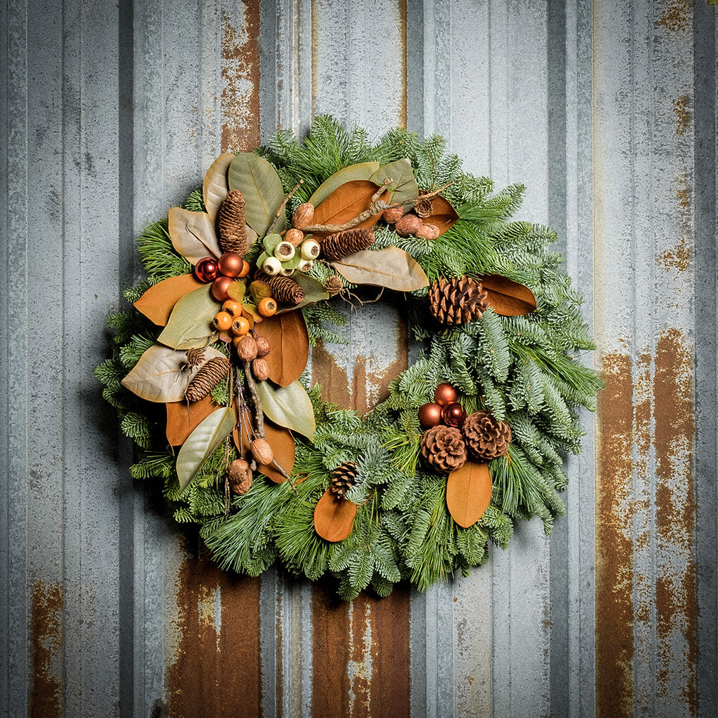 A wreath made of noble fir, white pine, and magnolia leaves with ponderosa and Austrian pinecones, copper balls, cream and orange seed pods, and branches of assorted nuts and cones on a rustic metal background.