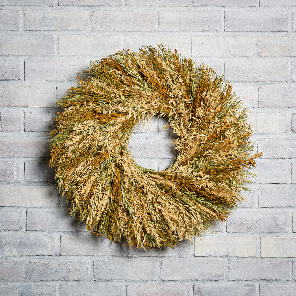 A wreath made of Sunkissed Aveena-oats, dijon Sudan grass, saffron flax, and natural wheat on a white brick background.