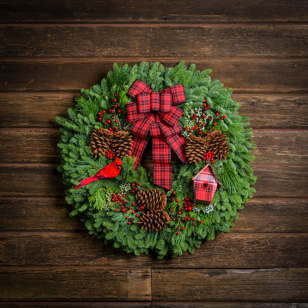 Holiday wreath made of noble fir, white pine, incense cedar, and juniper with ponderosa pine cones, red rosehip/mini pine cone clusters, faux berry clusters, red cardinal, a mini plaid birdhouse, and a red and black plaid bow hanging on a wooden wall