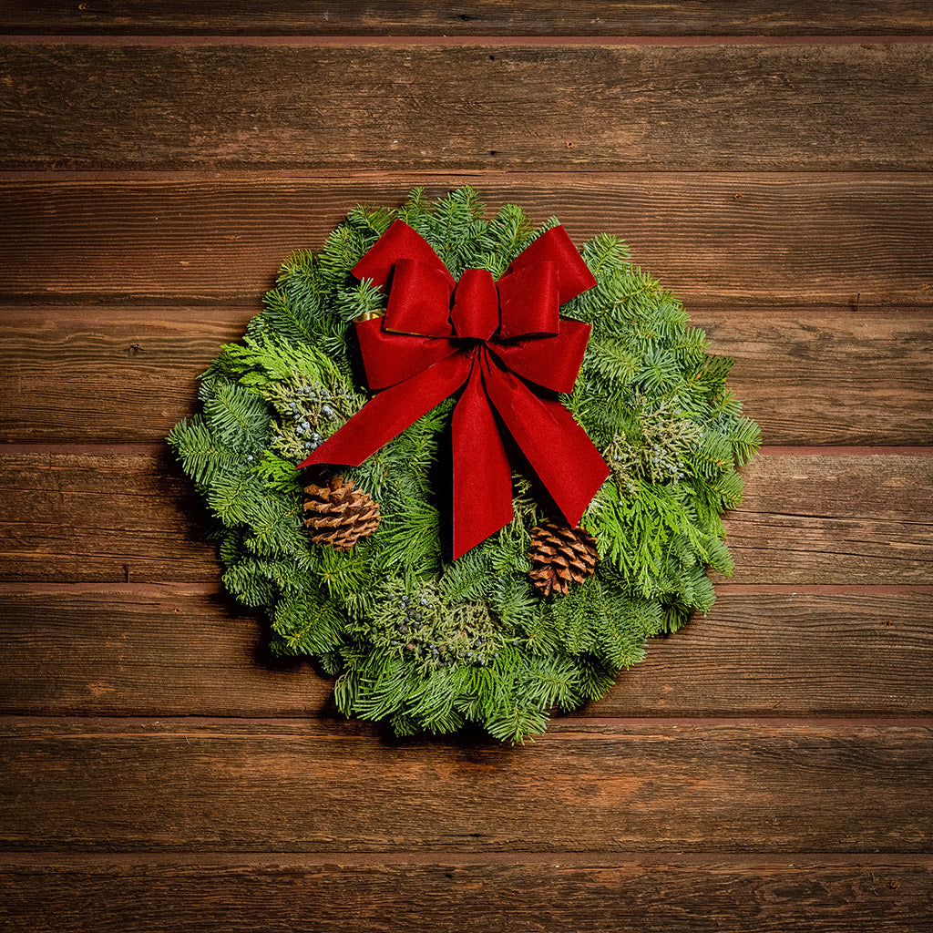 Christmas wreath made of fir, cedar, and juniper with pine cones and a red with gold back bow on a wood background.