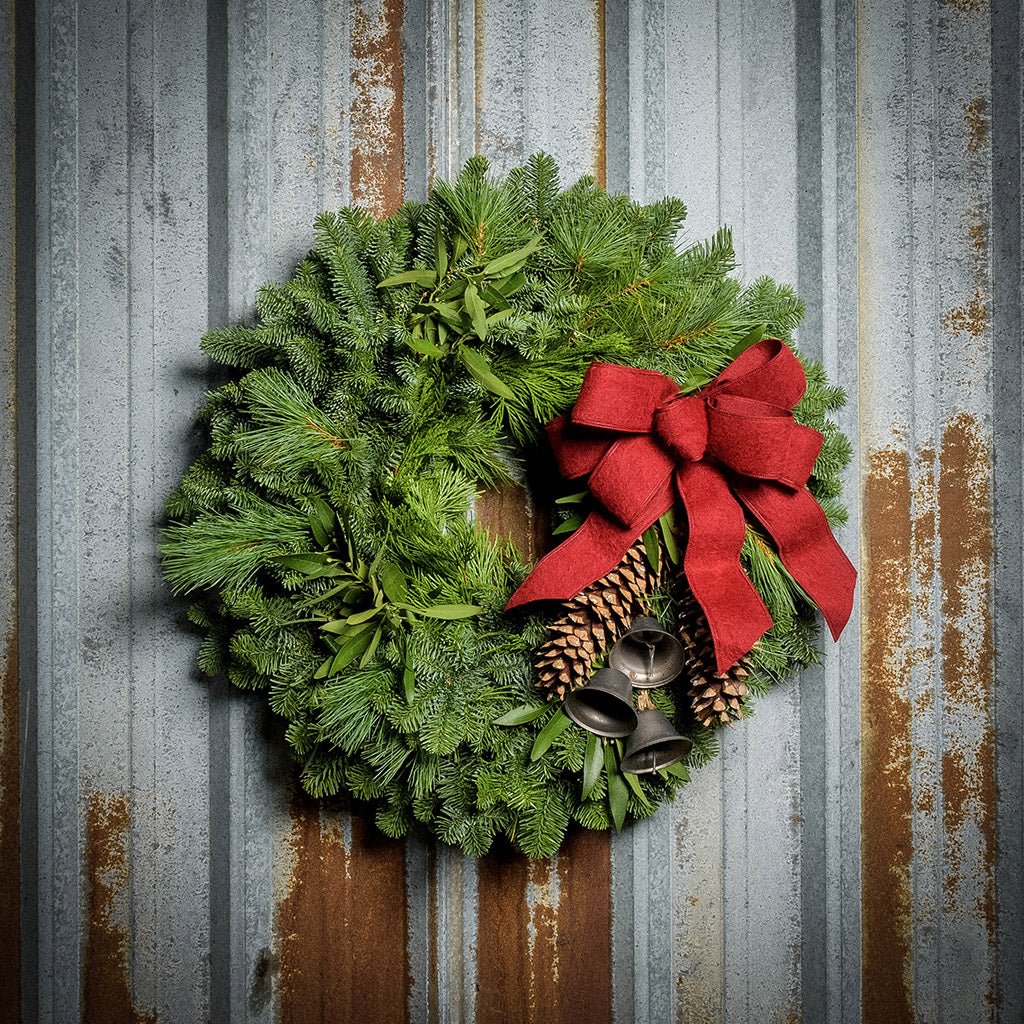 Holiday wreath made of noble fir, incense cedar, white pine, and California bay leaves with 6"- 8" white pine cones, burnished silver bells hanging from a jute string, and a red brushed linen bow on a rustic metal background.