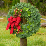 Holiday wreath with pine cones, faux autumn berries and a red linen bow on a base of noble, pine and salal greens on a post in field