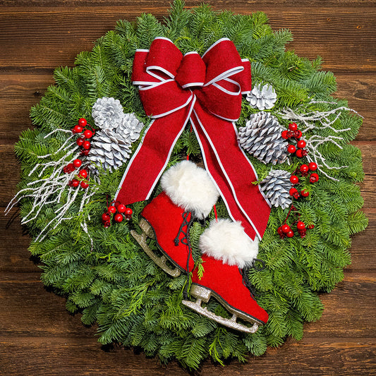 A festive Christmas wreath made of green pine branches, decorated with a large red fabric bow with white trim, red and white ice skates, silver pinecones, white twig-like ornaments, and red berries, displayed on a dark wood background.