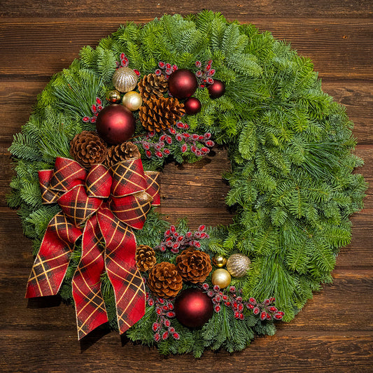 green pine wreath on a dark wood wall. The wreath is decorated with a large red plaid bow, pinecones, and red and gold ornaments.