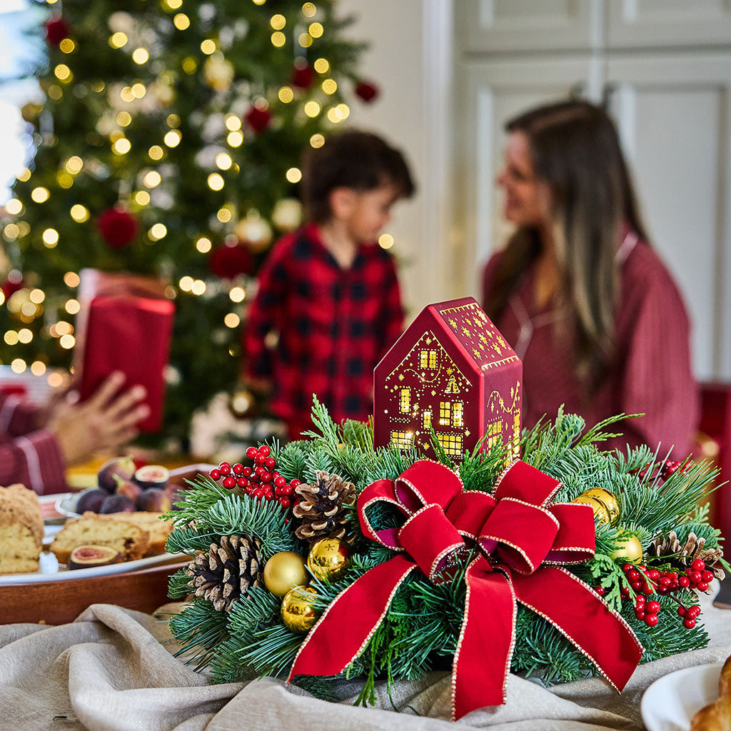 A festive Christmas centerpiece in the foreground with green pine branches, red ribbon, gold ornaments, and a small glowing red house ornament, set on a table in a warm holiday home with blurred people and a Christmas tree in the background.