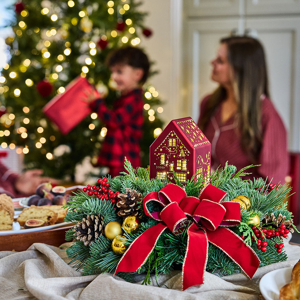 A festive Christmas centerpiece on a table, featuring green pine branches, a large red velvet bow with gold trim, red berries, pinecones, and a small red house ornament, with a blurred Christmas tree and two people in the background.