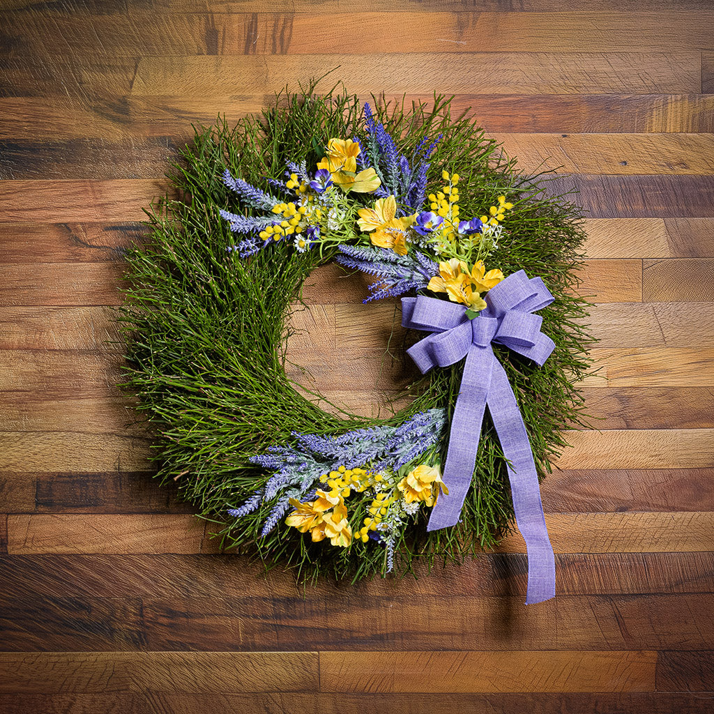 Green wreath with yellow and purple flowers and a purple ribbon on a wooden surface