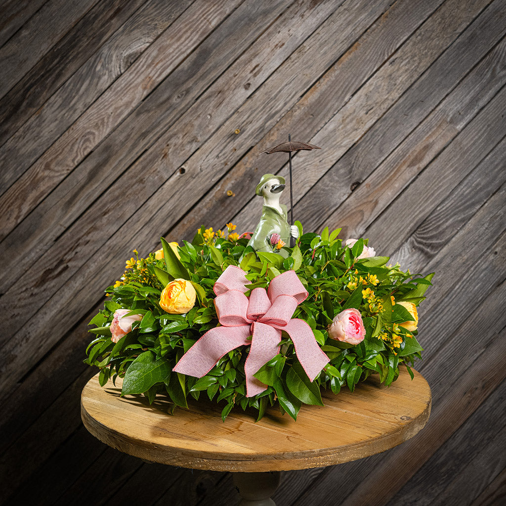 Fresh salal and green huckleberry with faux yellow and pink tulips, faux yellow wax flowers, mauve pink linen bows, and a white resin duck in green rain gear holding an umbrella