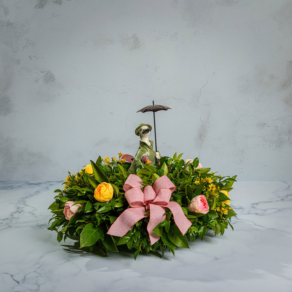 Fresh salal and green huckleberry with faux yellow and pink tulips, faux yellow wax flowers, mauve pink linen bows, and a white resin duck in green rain gear holding an umbrella