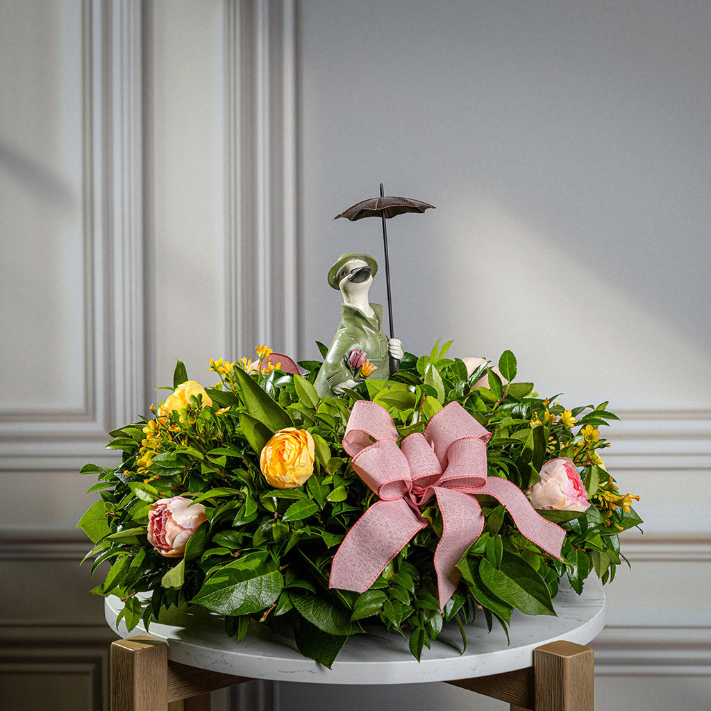 Fresh salal and green huckleberry with faux yellow and pink tulips, faux yellow wax flowers, mauve pink linen bows, and a white resin duck in green rain gear holding an umbrella