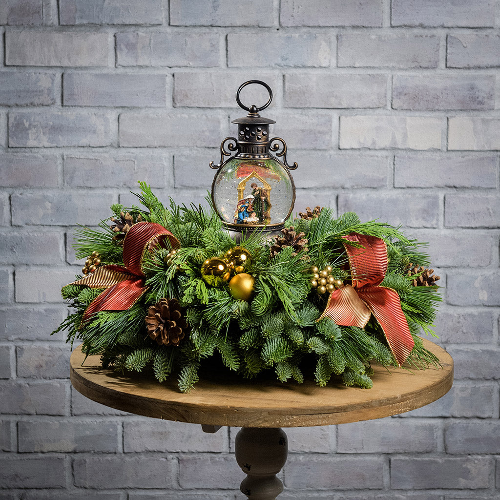 An evergreen arrangement of noble fir, cedar, and white pine with natural pinecones, gold ball ornaments, gold berries, gold bow tucks, and a nativity themed water globe