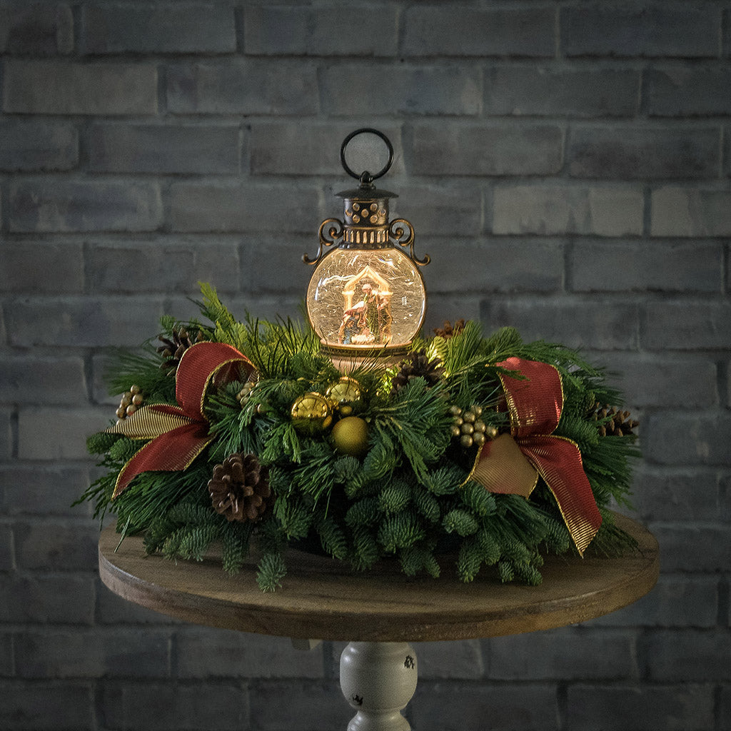 An evergreen arrangement of noble fir, cedar, and white pine with natural pinecones, gold ball ornaments, gold berries, gold bow tucks, and a nativity themed water globe