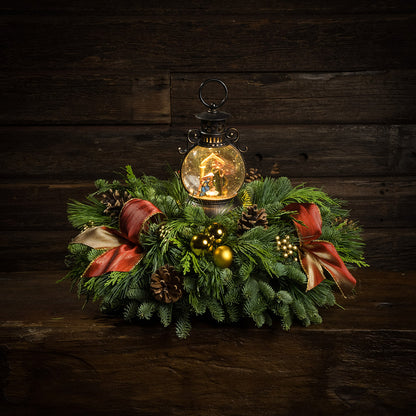 An evergreen arrangement of noble fir, cedar, and white pine with natural pinecones, gold ball ornaments, gold berries, gold bow tucks, and a nativity themed water globe