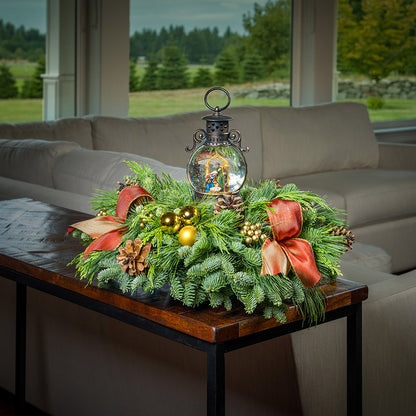 An evergreen arrangement of noble fir, cedar, and white pine with natural pinecones, gold ball ornaments, gold berries, gold bow tucks, and a nativity themed water globe