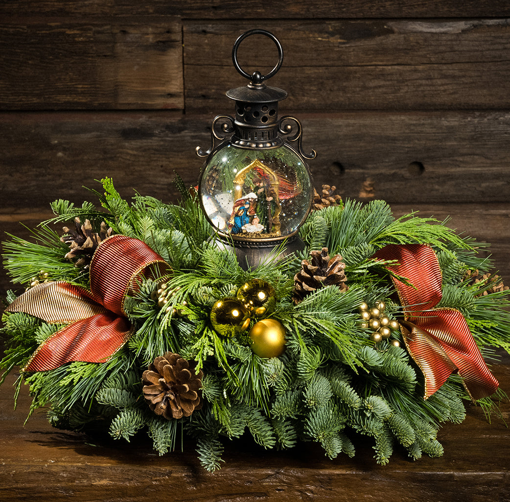 An evergreen arrangement of noble fir, cedar, and white pine with natural pinecones, gold ball ornaments, gold berries, gold bow tucks, and a nativity themed water globe