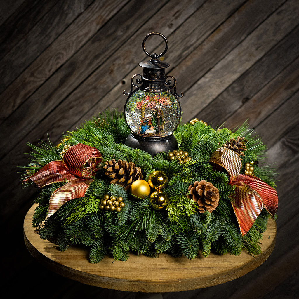 An evergreen arrangement of noble fir, cedar, and white pine with natural pinecones, gold ball ornaments, gold berries, gold bow tucks, and a nativity themed water globe