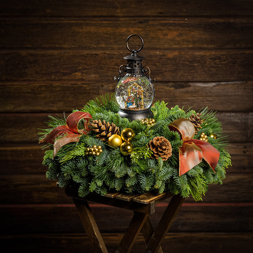 An evergreen arrangement of noble fir, cedar, and white pine with natural pinecones, gold ball ornaments, gold berries, gold bow tucks, and a nativity themed water globe