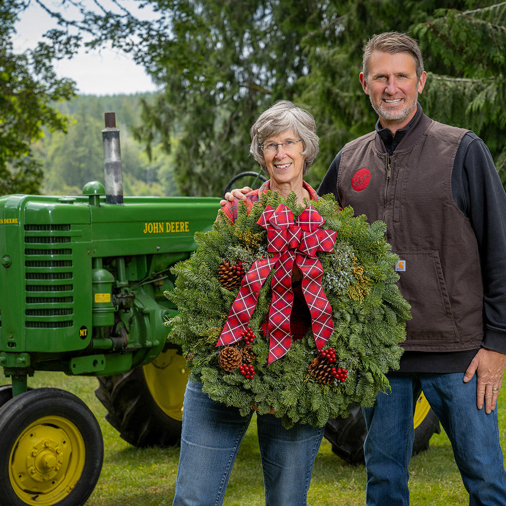 Two people holding a wreath with a red bow in front of a John Deere tractor.