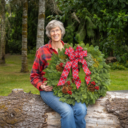 Woman holding a Christmas wreath with a red plaid bow, sitting on a log outdoors.