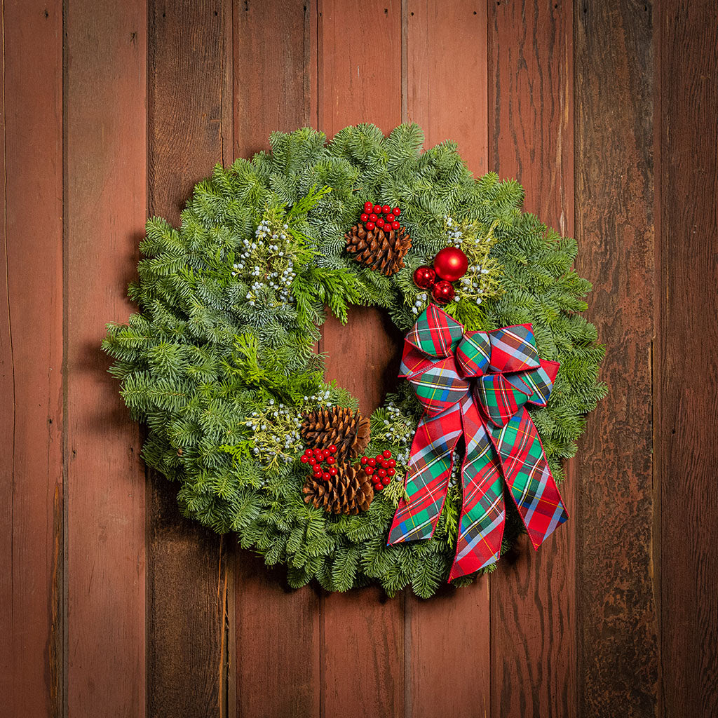 A festive Christmas wreath made of green pine branches, decorated with pinecones, red berries, white sprigs, and a large red and green plaid bow, hanging against a rustic brown wooden plank background.