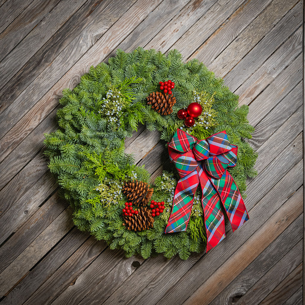 A festive evergreen Christmas wreath with pine cones, red berries, white flowers, and a large red and green plaid bow, displayed on a rustic wooden plank surface.