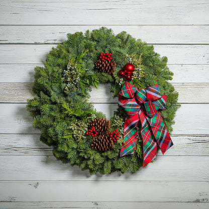 A festive Christmas wreath made of green pine branches, decorated with red berries, pinecones, small white berries, and a large red, green, and blue plaid bow, shown against a white wood plank background.