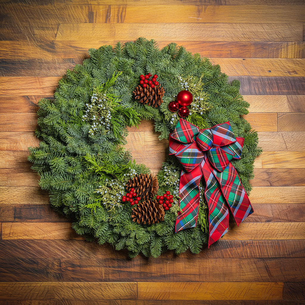 A green pine wreath decorated with red berries, pinecones, white berries, and a large red plaid bow, photographed from above on a wooden surface.
