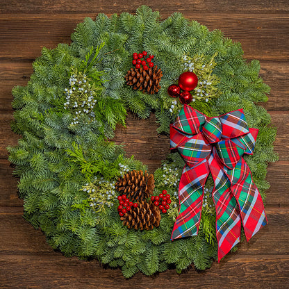 A festive Christmas wreath made of green pine branches, decorated with two pinecones, red berries, white berries, red ornaments, and a large red, green, and blue plaid bow, set against a dark wood background.