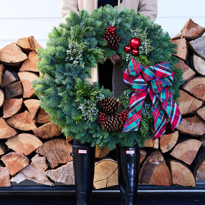 A large green evergreen Christmas wreath with pinecones, red ornaments, and a red plaid bow, held in front of a person wearing black rain boots and a beige coat, with stacked firewood in the background.